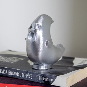 May include: A silver-colored 3D printed ghost figurine sitting on top of two books. The top book is black with white text that reads "Rasa Bugavicute-Pice" and the bottom book is red with white text that reads "Markizs de Sads Justine jeb Tikumitas nedienas JUMAVA".