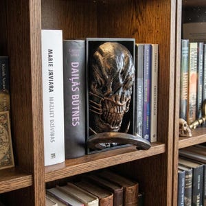 Könnte beinhalten: Ein Bücherregal aus Holz mit Büchern unterschiedlicher Größe und Farbe. Ein detailliertes, metallisches Alien-Kopf-Buchstütze wird zwischen zwei Büchern ausgestellt. Zusätzliche dekorative Schädel und eine Karte sind sichtbar.