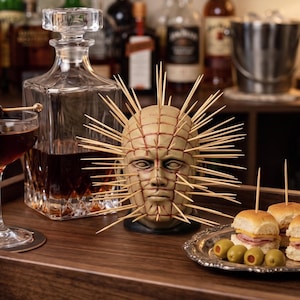 May include: A cocktail with a cherry garnish, a decorative head sculpture, a decanter, and appetizers on a wooden tray. Various bottles of liquor are in the background. The sculpture has a light brown color with wooden skewers.
