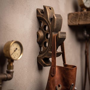 Pode incluir: Um gancho de parede em forma de engrenagem feito de metal em tom de bronze escuro com acabamento desgastado. Um avental de couro castanho com ferramentas de metal visíveis no bolso frontal está pendurado no gancho. Dois manómetros analógicos são visíveis.
