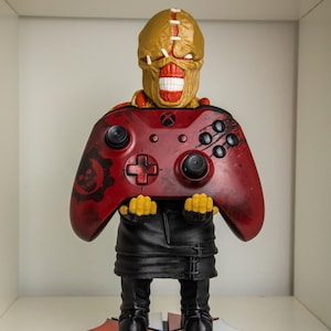 Puede incluir: Una figura de un monstruo sosteniendo un mando de Xbox rojo. El monstruo tiene una cabeza bronceada con puntadas y la boca muy abierta. El mando tiene botones negros y un logotipo de Gears of War. La figura se apoya en una base negra, blanca y roja.
