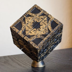 May include: A gold and black cube puzzle with intricate carvings and a starburst design in the center. The cube is on a gold stand.