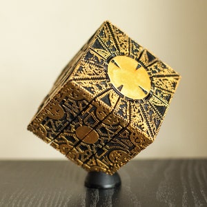 Puede incluir: Una caja de rompecabezas en forma de cubo de color dorado y negro con diseños intrincados y un círculo central dorado. La caja está sobre un soporte negro.
