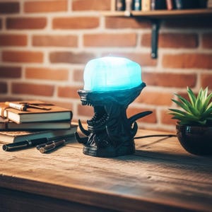 May include: A black lamp shaped like the head of a xenomorph creature with a translucent blue dome top. The lamp features a detailed jaw with visible teeth and pointed protrusions, emitting a soft blue light from the upper portion of the head.