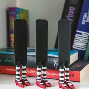 A black bookmark shaped like the Wicked Witch of the West's legs, complete with red shoes and striped stockings, sticking out from  books on a shelf.