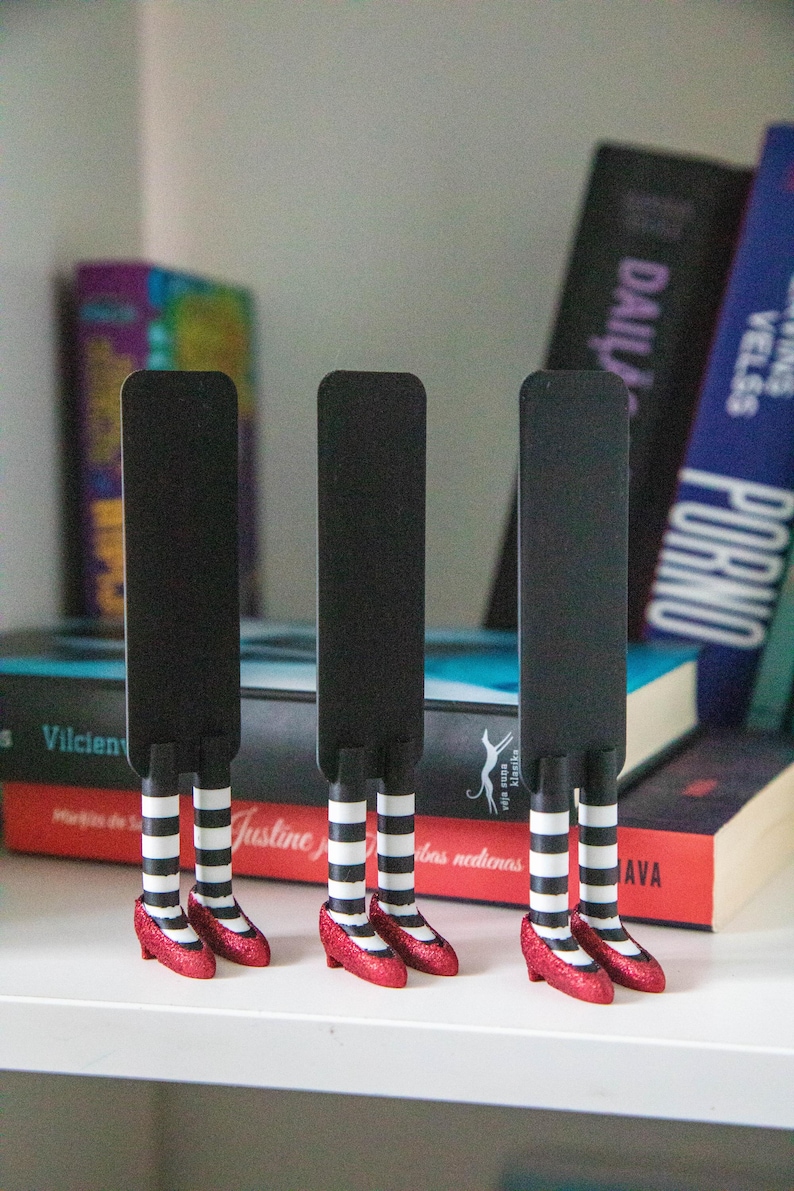 A black bookmark shaped like the Wicked Witch of the West's legs, complete with red shoes and striped stockings, sticking out from  books on a shelf.