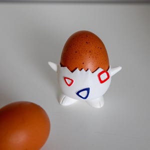 Puede incluir: Un porta huevos blanco con forma de personaje de dibujos animados con detalles rojos, azules y blancos. El porta huevos contiene un huevo marrón.