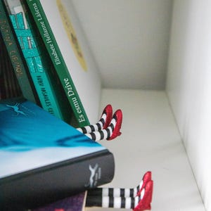 A black bookmark shaped like the Wicked Witch of the West's legs, complete with red shoes and striped stockings, sticking out from  books on a shelf.