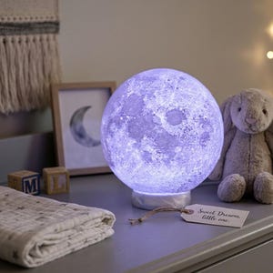 Könnte beinhalten: Eine kugelförmige Mondlampe, die ein violettes Licht ausstrahlt und auf einem weißen Sockel ruht. Ein kleines rechteckiges Etikett am Sockel trägt den aufgedruckten Text "Sweet Dreams, little one." Die Lampe hat eine texturierte Oberfläche.
