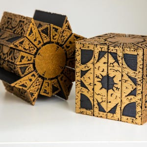 May include: Two intricately designed puzzle boxes. One is a cube with geometric patterns and black accents. The other is a complex, multi-faceted shape with a central circular element and black panels. Both are in shades of gold and brown.