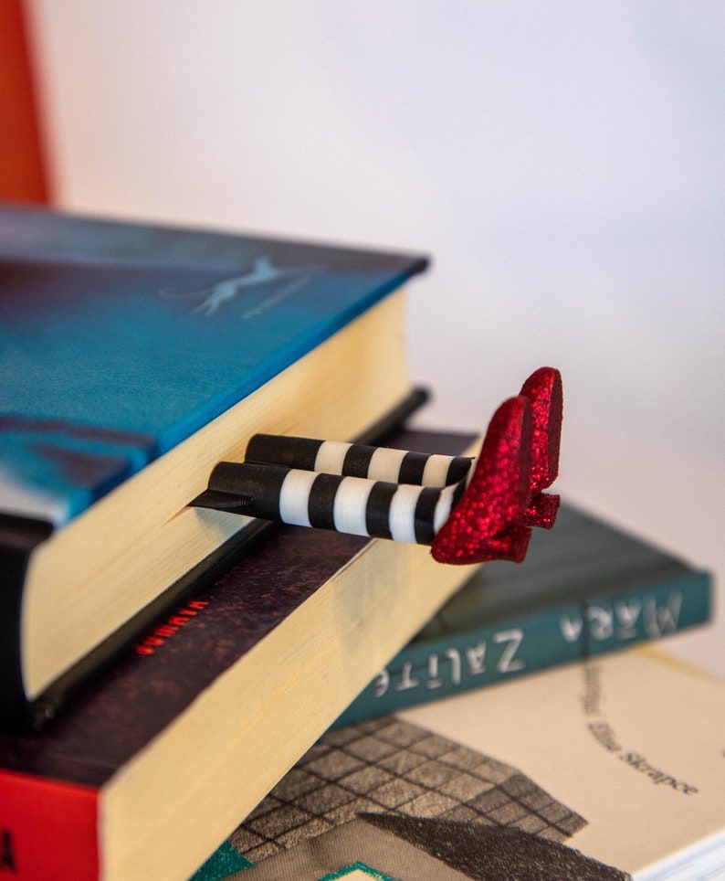 A black bookmark shaped like the Wicked Witch of the West's legs, complete with red shoes and striped stockings, sticking out from  books on a shelf.