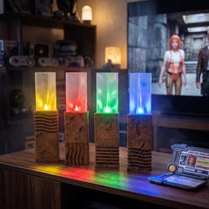 Peut inclure: Quatre piliers décoratifs lumineux avec une base imitation bois et des dessus rectangulaires transparents. Chaque pilier émet une couleur différente : jaune, rouge, vert et bleu. Les lumières sont disposées en rangée sur une surface en bois, avec un arc-en-ciel de couleurs projeté.