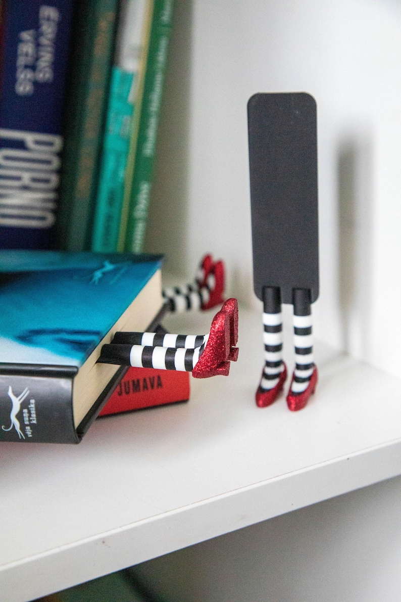 A black bookmark shaped like the Wicked Witch of the West's legs, complete with red shoes and striped stockings, sticking out from  books on a shelf.