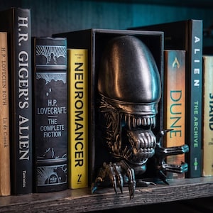 May include: A dark metallic Xenomorph creature sculpture positioned as a bookend between several books, including titles like H.R. Giger's Alien, H.P. Lovecraft: The Complete Fiction, Neuromancer, Dune, Alien: The Archive, and The Stand.
