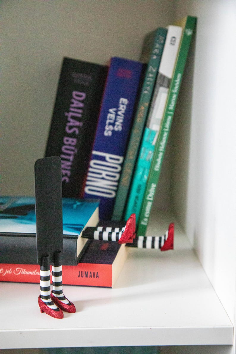 A black bookmark shaped like the Wicked Witch of the West's legs, complete with red shoes and striped stockings, sticking out from  books on a shelf.