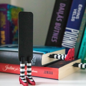 A black bookmark shaped like the Wicked Witch of the West's legs, complete with red shoes and striped stockings, sticking out from  books on a shelf.
