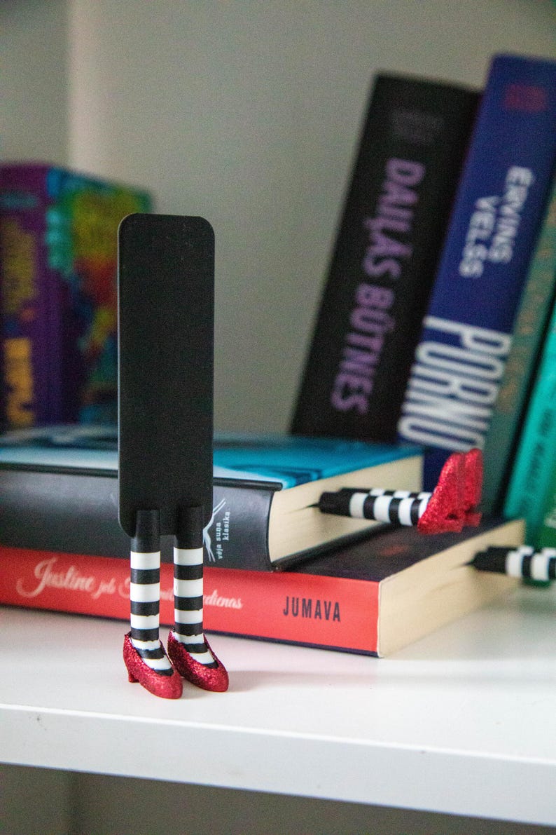 A black bookmark shaped like the Wicked Witch of the West's legs, complete with red shoes and striped stockings, sticking out from  books on a shelf.