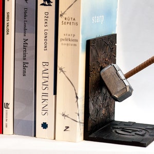 Puede incluir: Un par de sujetalibros con forma de martillo de Thor, Mjolnir, con un mango de madera marrón y una cabeza de metal plateado. Los sujetalibros están hechos de un material marrón oscuro con una superficie texturizada. Los sujetalibros sostienen una pila de libros.