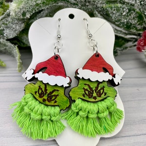 May include: A pair of Grinch-themed earrings. Each earring features a green Grinch face with a red Santa hat and a green tassel. The earrings are made of wood and yarn, and hang from silver-colored hooks.