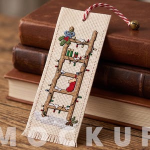 Può includere: Un segnalibro rettangolare in tessuto con un disegno a punto croce di una scala in legno decorata con luci natalizie, una calza rossa e del verde. Il segnalibro presenta un bordo sfrangiato e un cordino rosso e bianco con un campanellino.