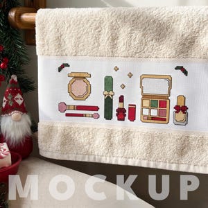 Può includere: Un asciugamano in spugna beige con un motivo a punto croce di articoli per il trucco, tra cui una cipria compatta, pennelli, un rossetto, una palette di ombretti e uno smalto, decorato con piccole foglie di agrifoglio e croci dorate.