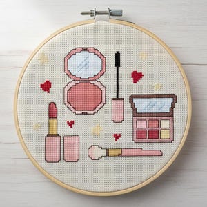 Può includere: Ricamo a punto croce in un telaio di legno, raffigurante articoli per il trucco. Il design include un compatto con specchio, mascara, rossetto, un pennello per il trucco e una palette di ombretti. La tavolozza dei colori è principalmente rosa e beige, con accenti a cuore rosso.