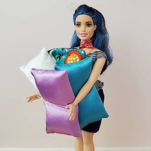 May include: A blue-haired doll holds a stack of colorful pillows. The pillows are made of satin and have different patterns. The doll is wearing a striped shirt and a red bandana.