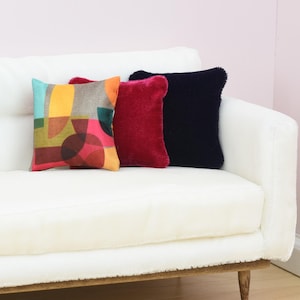 May include: Three decorative throw pillows on a white couch. The pillows are a geometric pattern with red, yellow, blue, and green colors, a solid red velvet pillow, and a solid navy blue velvet pillow.