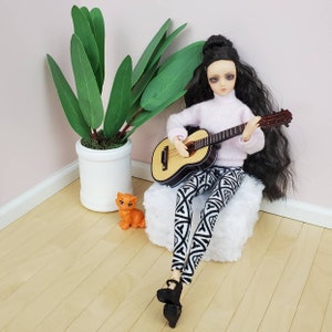 K&ouml;nnte beinhalten: Eine Puppe mit langen schwarzen Haaren spielt eine Miniatur-Akustikgitarre. Die Puppe tr&auml;gt einen hellrosa Pullover, eine Hose mit schwarz-wei&szlig;em Muster und schwarze Plateauschuhe. Eine kleine orangefarbene Katzenfigur und eine Topfpflanze sind ebenfalls zu sehen.