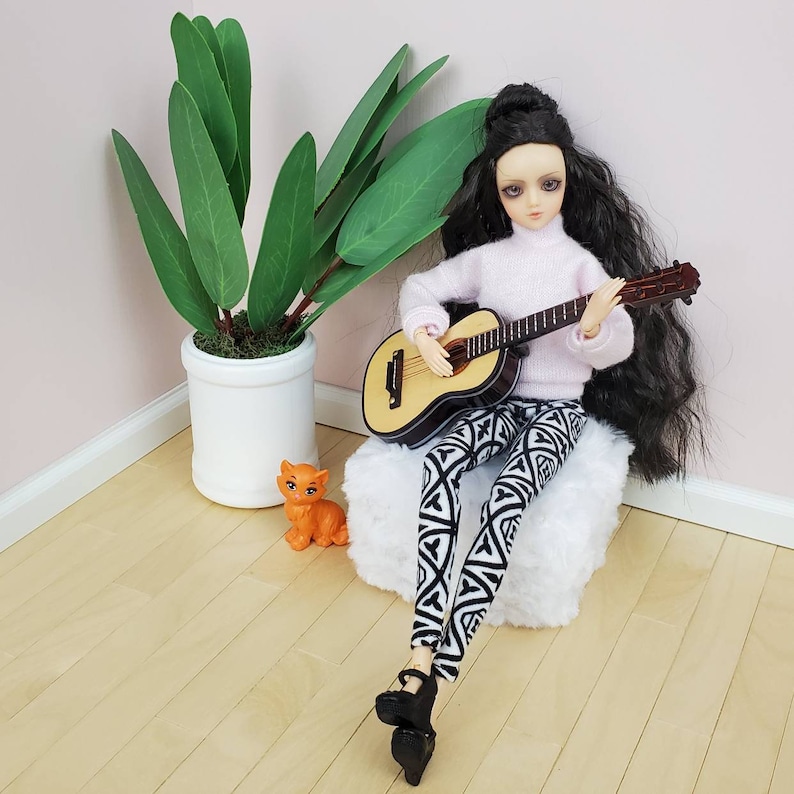 K&ouml;nnte beinhalten: Eine Puppe mit langen schwarzen Haaren spielt eine Miniatur-Akustikgitarre. Die Puppe tr&auml;gt einen hellrosa Pullover, eine Hose mit schwarz-wei&szlig;em Muster und schwarze Plateauschuhe. Eine kleine orangefarbene Katzenfigur und eine Topfpflanze sind ebenfalls zu sehen.