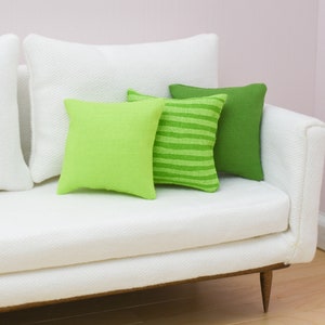 May include: A white couch with three green throw pillows. The pillows are different shades of green and have different textures. The couch has a wooden frame and legs.