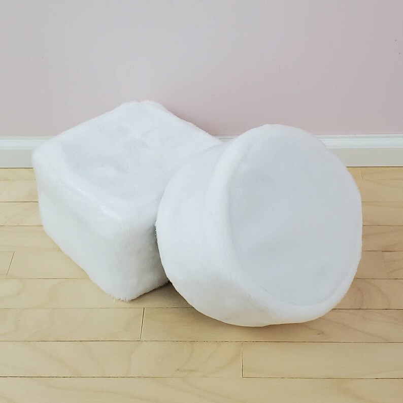 K&ouml;nnte beinhalten: Zwei wei&szlig;e, pl&uuml;schige Ottomanen. Einer ist w&uuml;rfelf&ouml;rmig, der andere zylindrisch. Beide bestehen aus einem weichen, pelzigen Material. Sie stehen auf einem hellen Holzboden, mit einer blassrosa Wand im Hintergrund.