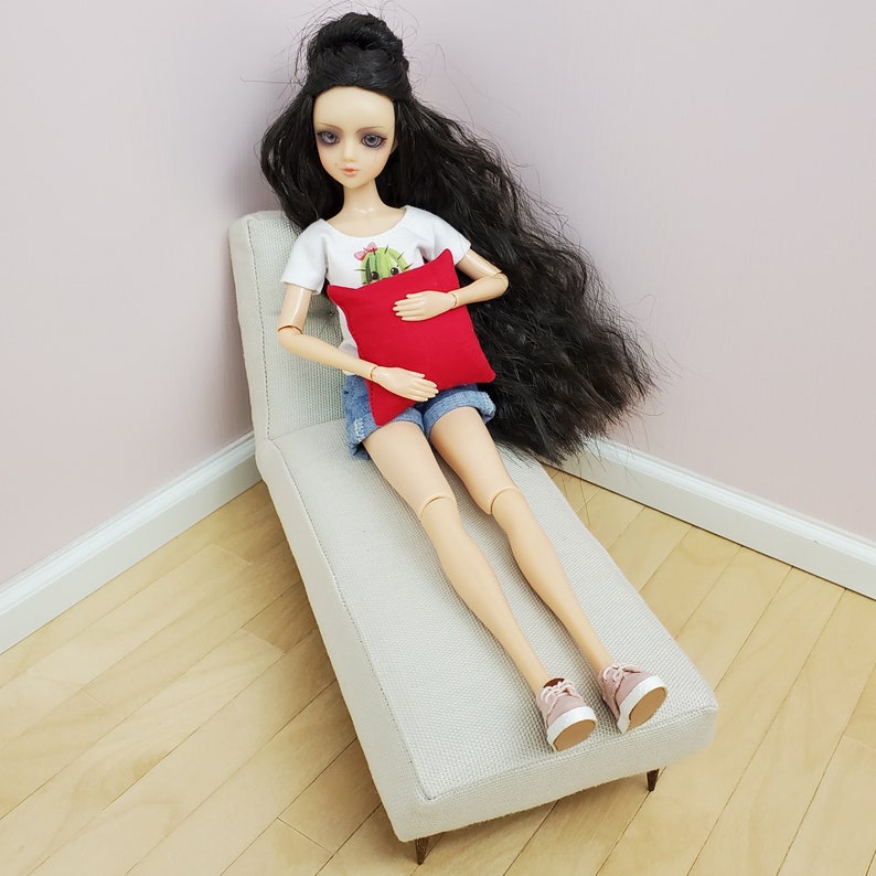 May include: A doll with long black hair is sitting on a white chaise lounge. She is wearing a white shirt with a cactus design, denim shorts, and pink sneakers. She is holding a red pillow.