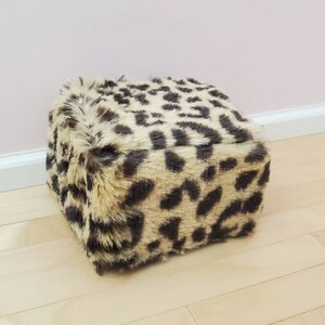 K&ouml;nnte beinhalten: Ein quadratischer Hocker mit einem Leopardenmuster aus Kunstfell. Der Hocker hat einen beigefarbenen Hintergrund mit dunkelbraunen Flecken. Das Pl&uuml;schmaterial verleiht ihm ein weiches, strukturiertes Aussehen. Er steht auf einem hellen Holzboden.
