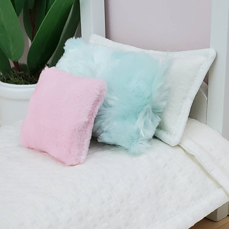 May include: Two fluffy pillows, one light pink and one light blue, are placed on a white bed with a white pillow behind them.