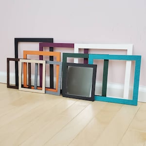 May include: A collection of 10 different colored picture frames, all in various sizes, are arranged in a row on a light wood floor. The frames are made of wood and have a variety of finishes, including black, white, teal, orange, and purple.