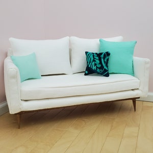 May include: A white sofa with a white textured fabric upholstery. The sofa has three pillows, two are light blue and one is dark blue with a teal floral pattern.