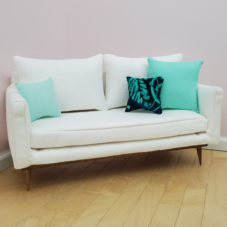 May include: A white sofa with a white textured fabric upholstery. The sofa has three pillows, two are light blue and one is dark blue with a teal floral pattern.