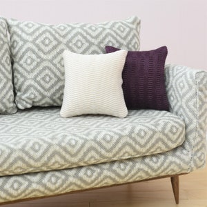 May include: A gray couch with a geometric pattern, featuring three pillows. Two pillows are gray with a geometric pattern, and one pillow is white with a wavy pattern. The couch has wooden legs.