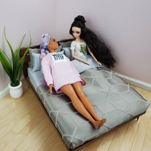 May include: Two dolls are lying on a gray bed with a hexagonal pattern. The doll on the left is wearing a pink sweatshirt with the text "Dream Often" printed on it. The doll on the right is wearing a white shirt with a cactus print. The bed has a wooden frame and a gray headboard.
