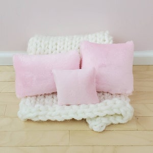 May include: Three pink fluffy pillows of different sizes are arranged on a white knitted blanket. The blanket is on a light wood floor.