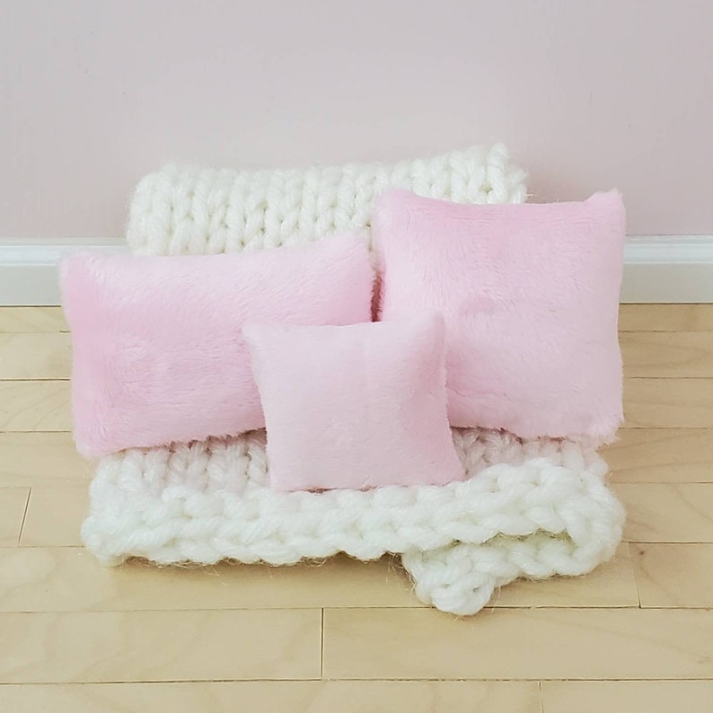 May include: Three pink fluffy pillows of different sizes are arranged on a white knitted blanket. The blanket is on a light wood floor.