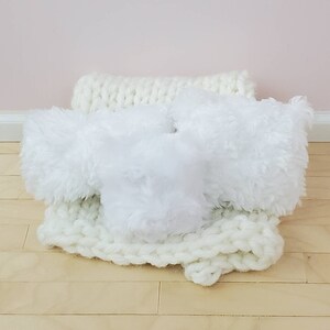 May include: Three white fluffy cushions on a white knitted throw. The cushions are soft and plush, and the throw is made of a thick, cosy yarn.