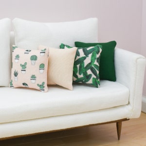 May include: A white couch with four throw pillows. Two pillows are green with a leafy pattern, one is a solid green, and one is pink with a cactus pattern.
