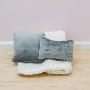 May include: Two grey faux fur pillows and a white knitted throw blanket. The pillows are rectangular and square shaped.