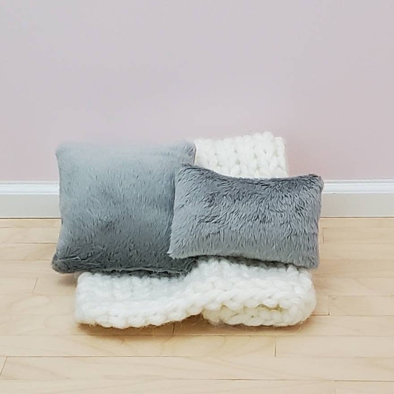 May include: Two grey faux fur pillows and a white knitted throw blanket. The pillows are rectangular and square shaped.