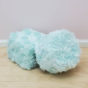 K&ouml;nnte beinhalten: Zwei flauschige, hellblaue dekorative Poufs. Die Poufs haben eine weiche, strukturierte Oberfl&auml;che mit einem rosenartigen Muster. Sie stehen auf einem hellen Holzboden, mit einer blassrosa Wand im Hintergrund.
