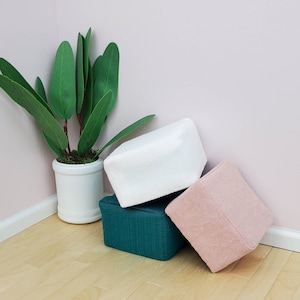 May include: Three fabric storage cubes in different colors and textures. The cubes are stacked on top of each other. The top cube is white, the middle cube is teal, and the bottom cube is pink.