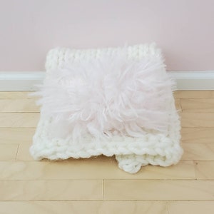 May include: A white knitted cushion with a fluffy pink insert. The cushion is soft and plush, ideal for adding a touch of comfort and style to any room.