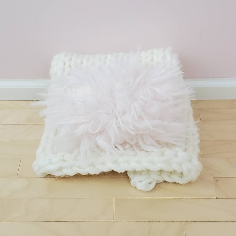May include: A white knitted cushion with a fluffy pink insert. The cushion is soft and plush, ideal for adding a touch of comfort and style to any room.